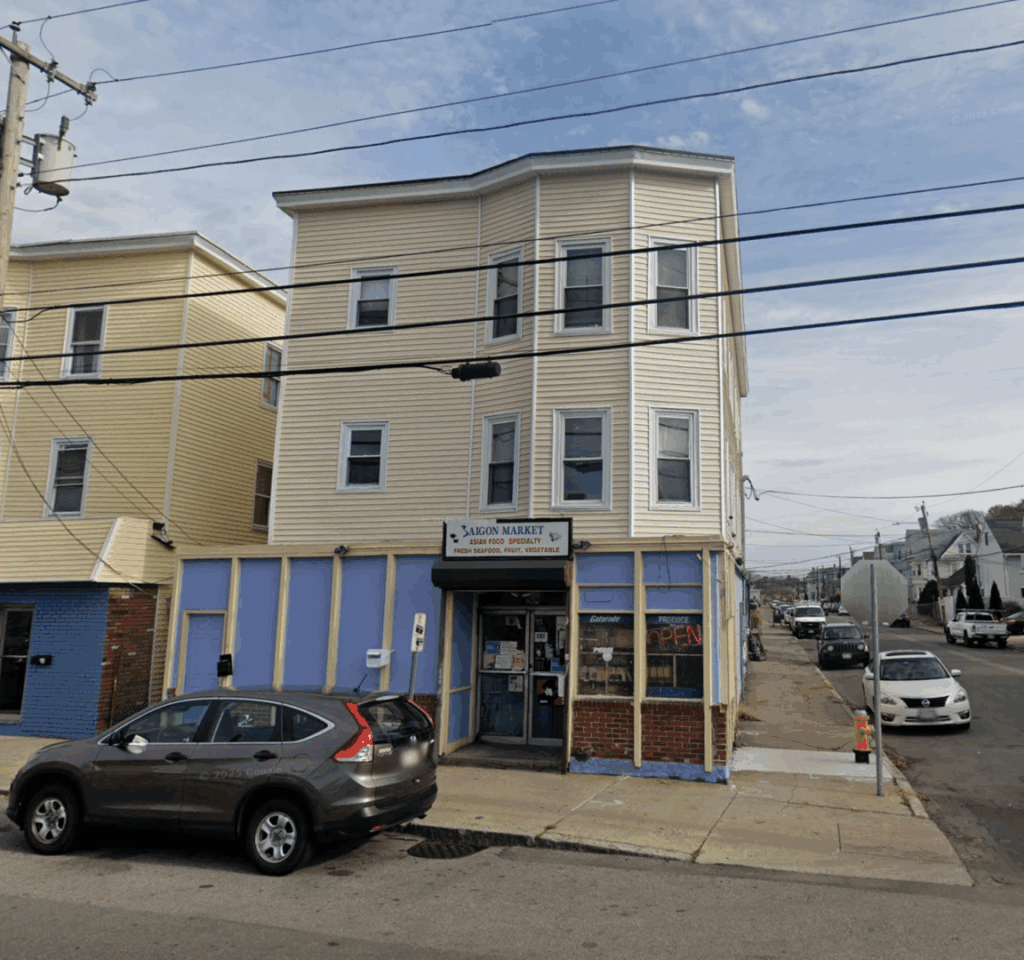 an image of a small market with blue paneled walls. It has a sign above the door reading "Saigon Market Asian Food Specialty: Fresh Seafood, Fruit, Vegetable. On top of the market is a small apartment with white siding.